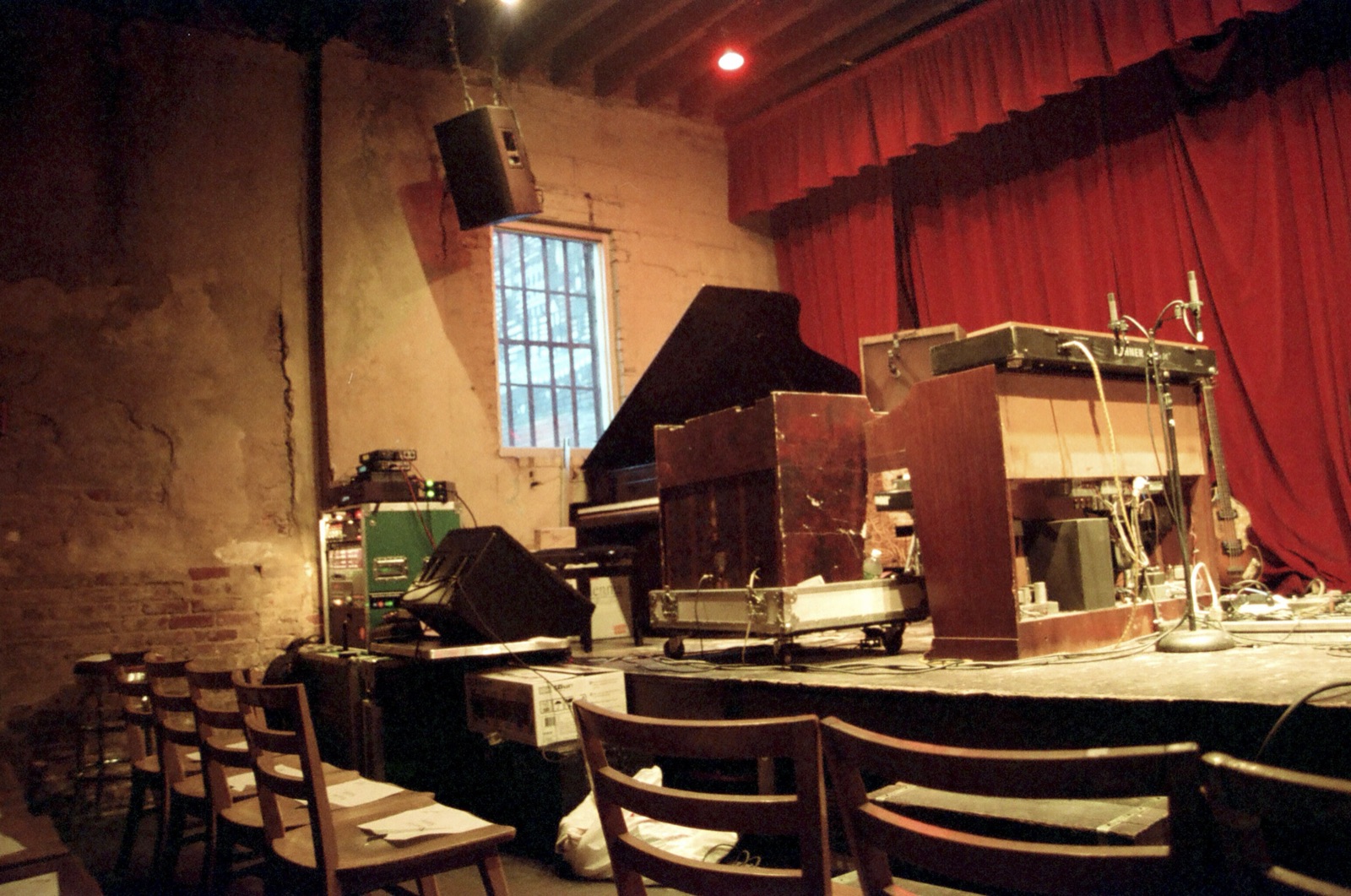 The inside of a small brick club, rows of wooden chairs facing a stage crowded with a piano, organ and gear under a red curtain.